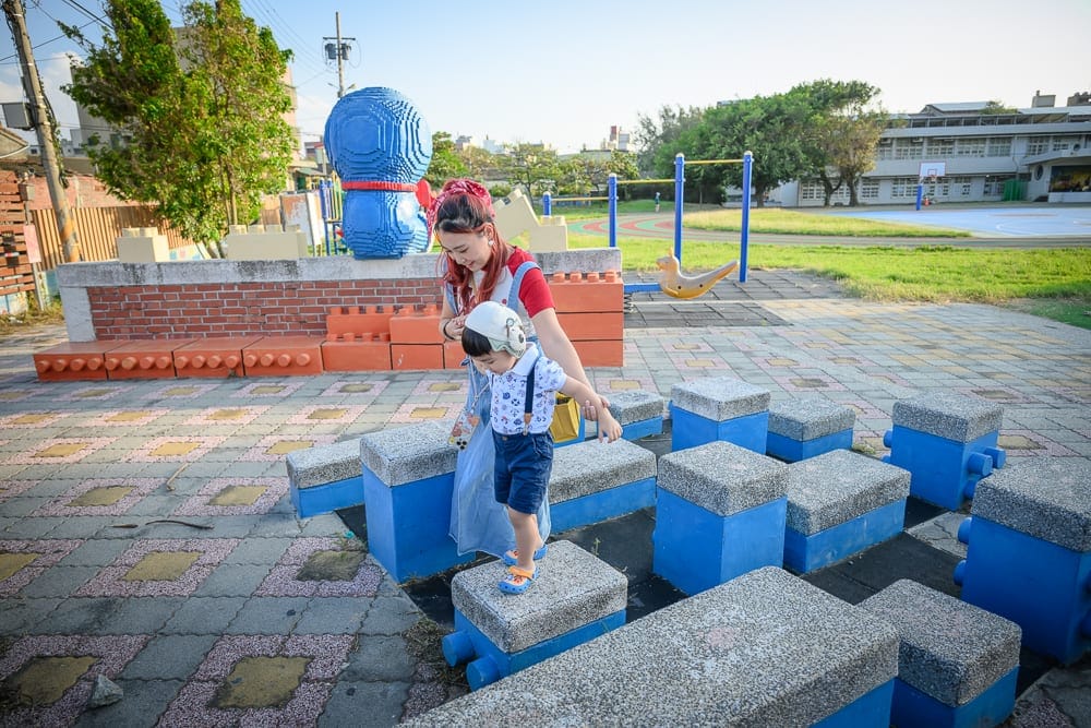 雲林麥寮國小積木公園｜戶外公園，婦人牽著幼童，積木造型座椅，彩色地磚，藍色大型機器人雕塑，單杠，操場。