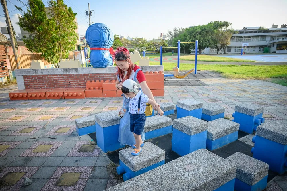雲林麥寮國小積木公園完整指南|親子遊樂設施與積木造景玩法 9 雲林麥寮國小積木公園|戶外公園,婦人牽著幼童,積木造型座椅,彩色地磚,藍色大型機器人雕塑,單杠,操場。