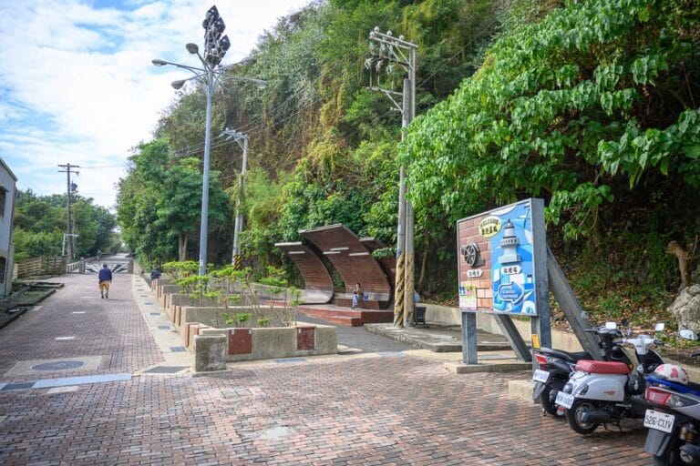 高雄旗津燈塔｜街道,木造座椅,告示牌,機車,路燈,山壁,綠樹,行人。磚造路面旁設有半圓弧型木椅與藍色觀光告示牌，右