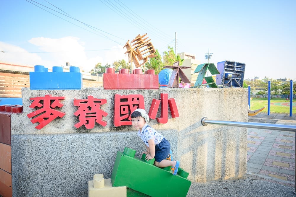 雲林麥寮國小積木公園｜幼童、國小校門口、巨型積木、紅字招牌、廣場、欄杆。戶外日照充足，淺色石牆上方裝飾藍紅兩色積木與木造文