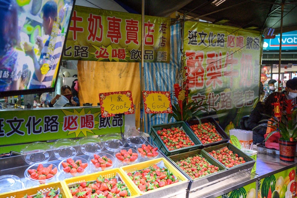 嘉義市文化路觀光夜市完整攻略|代表小吃與逛街指南 6 嘉義文化路夜市|夜市攤販,草莓,招牌,塑膠盒,螢幕,黃燈,行人。黃色綠色招牌下擺滿草莓塑膠盒,上方懸掛宣傳螢幕與布條