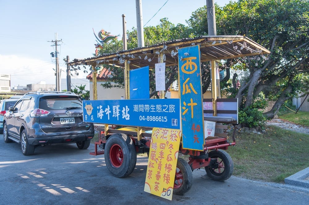 彰化海空步道｜街道、攤車、汽車、招牌、樹木，路旁停放一輛改裝的藍黃色餐車，車身懸掛海牛驛站與西瓜汁招牌，左側停著黑