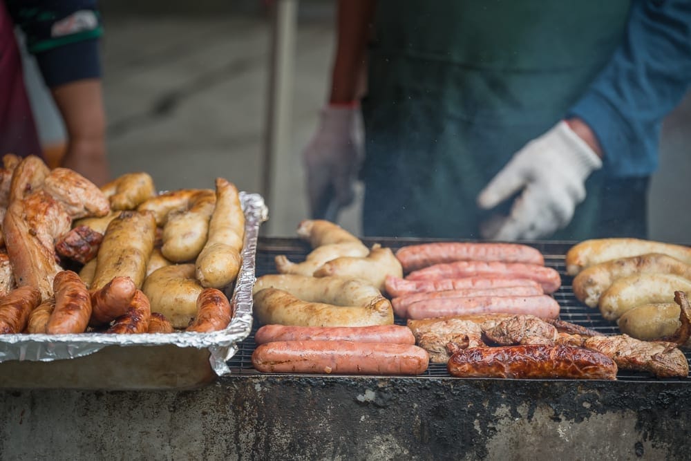 高雄寶來二集團櫻花公園｜料理, 香腸, 米腸, 烤肉架, 攤位。攤販戴著白手套與綠圍裙，在烤架前翻動各種肉腸。鋁箔紙盤內堆滿