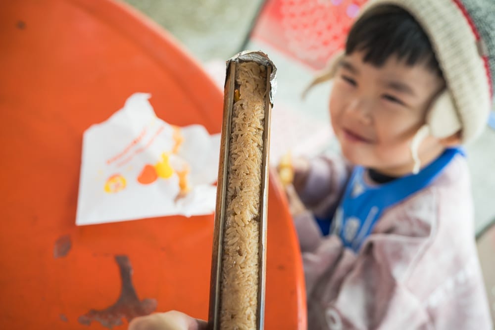 高雄寶來二集團櫻花公園｜竹筒飯，料理，兒童，圓桌，白色紙袋。剖開的竹筒內裝滿長形米飯，表面附著竹膜。右側幼童戴著毛帽與耳罩，