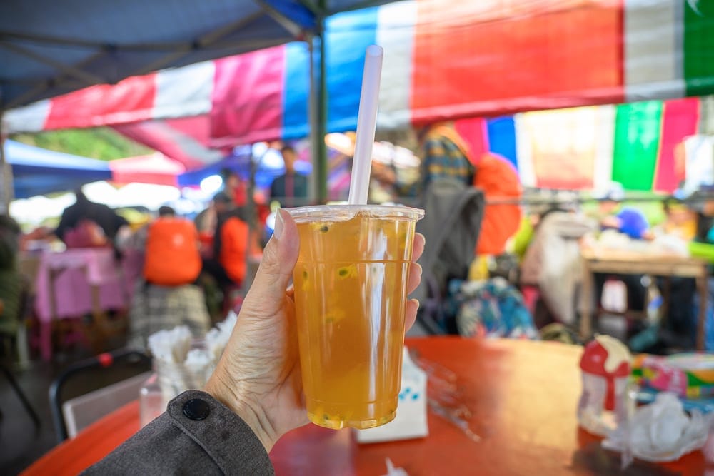 高雄寶來二集團櫻花公園｜手、塑膠杯、百香果綠茶、吸管、紅桌子、戶外市集、彩色遮陽棚、人群。手持透明塑膠杯，內裝黃色茶飲與百香