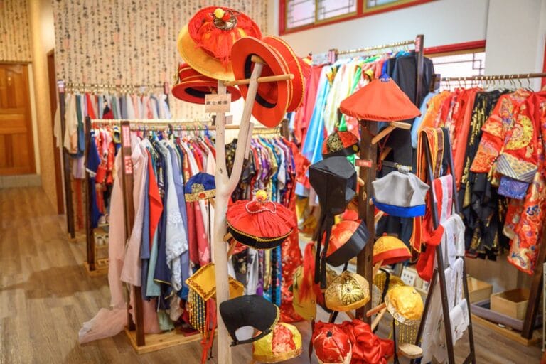 台南正統鹿耳門聖母廟漢服體驗｜室內、服飾店、衣服、帽子、衣架。架上掛滿色彩鮮豔的古裝服飾，木製立架上堆疊多款紅黑相間的中式官帽與圓