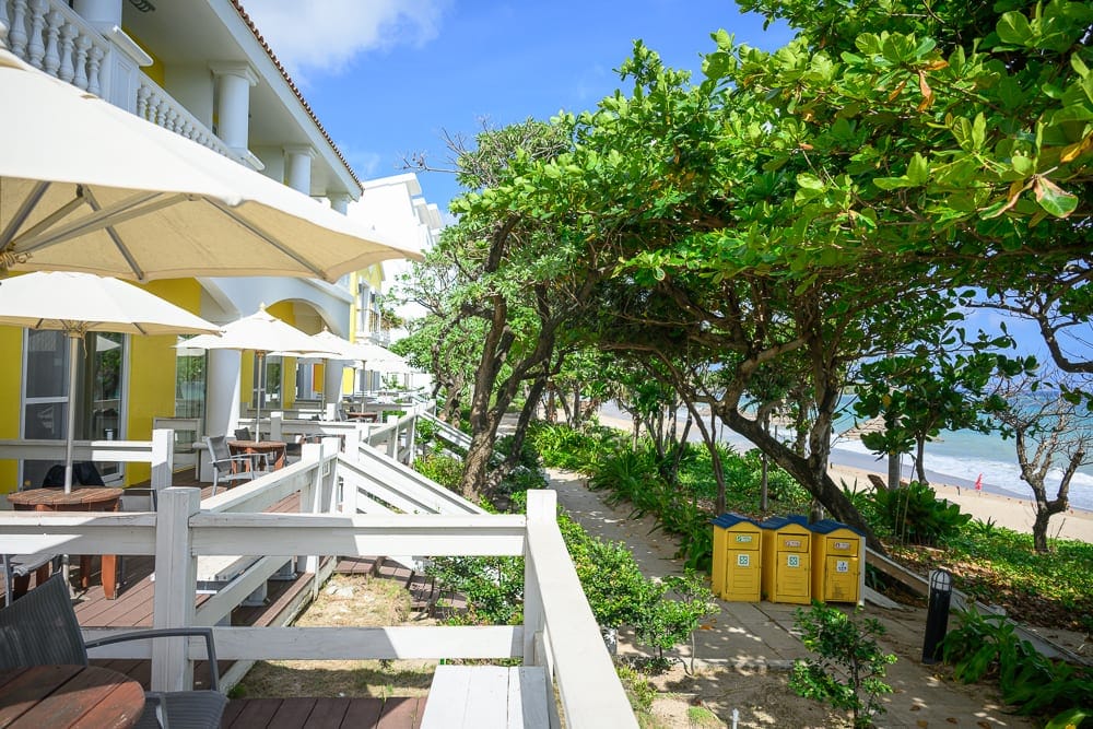 屏東夏都渡假飯店｜白牆建築、黃色柱子、陽傘、木造露台、綠樹、沙灘、海洋。海濱度假村露台，白色欄杆外並排幾支白色遮陽傘，