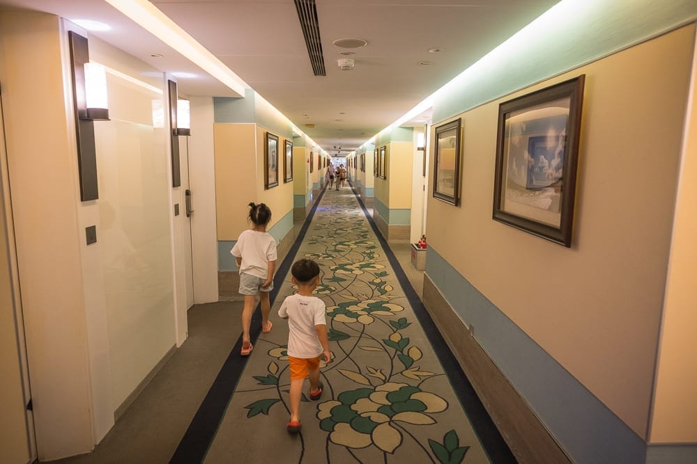 屏東夏都渡假飯店｜室內、走廊、小孩、地毯、壁畫，兩名幼童走在鋪有花卉圖騰地毯的長廊上，兩側米色牆面懸掛多幅框畫與壁燈，