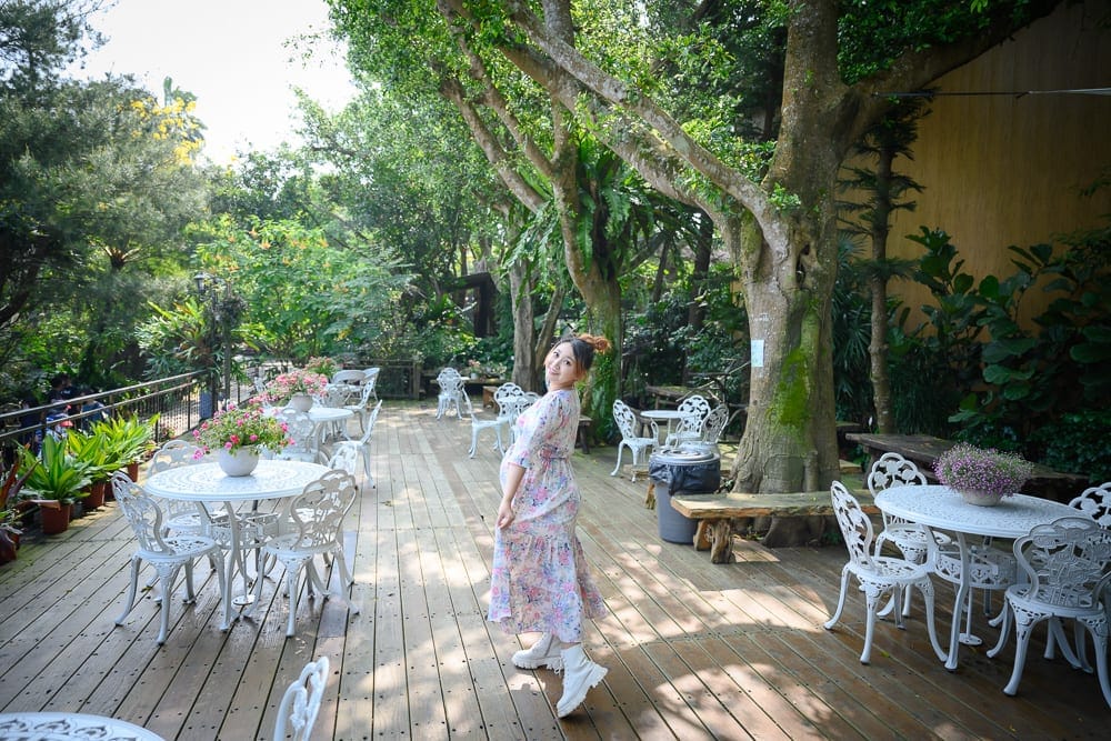 苗栗花露農場｜戶外木棧道平台、白色圓桌、鐵藝靠背椅、茂密綠樹、盆栽花卉、孕婦。女子身穿碎花洋裝與白靴，側身站立回眸