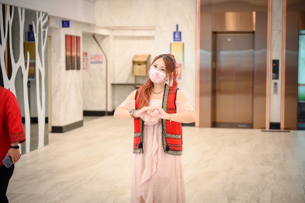 台中統一渡假村 谷關溫泉養生會館完整攻略|親子設施與溫泉泡湯全解析 12 台中谷關統一渡假村|室內、女性、電梯、長洋裝、背心,女子戴粉紅口罩雙手比心,身處大理石地坪走廊,背景有金色電梯門及立柱。