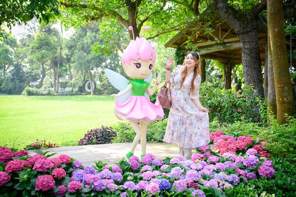 苗栗花露農場｜戶外草地、木平台、粉紅精靈立牌、長裙女子、花叢、樹木、涼亭，女子身穿碎花洋裝並揮手，立牌造型具備翅膀