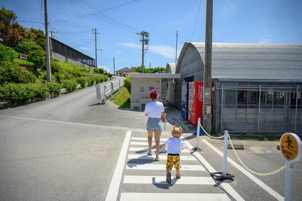 日本沖繩釣魚與探索冒險樂園完整攻略|親子體驗玩法解析 6 日本沖繩釣魚與探索冒險樂園|街道、斑馬線、電線桿、自動販賣機、鐵皮屋、坡道、行人。大人與戴帽孩童正走過斑馬線,前方為上坡柏油路,