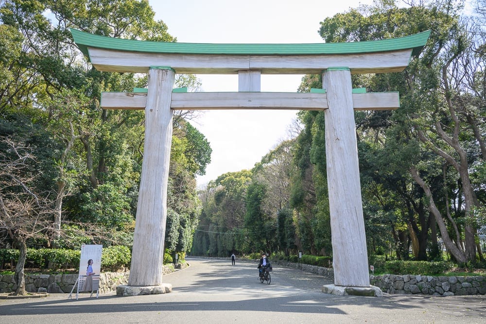 日本福岡-護國神社｜鳥居、林道、單車騎士、看板、樹木。白色巨大木造鳥居橫跨柏油路面，兩側立著石牆與茂密樹林，遠處道路中央