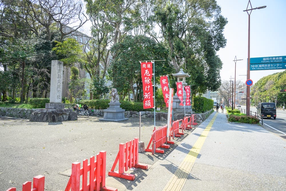 日本福岡-護國神社｜街道、石碑、石燈籠、紅色旗幟、行人道、路燈、樹木、車輛，戶外白天光線照射著馬路與人行道，遠方有指標與
