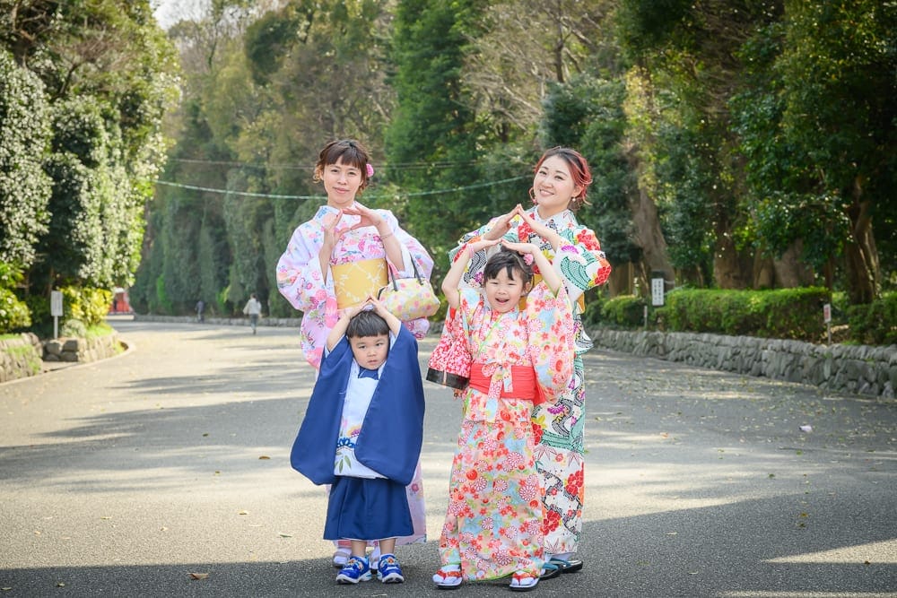 日本福岡-護國神社｜街道、和服、兩位女性、兩個小孩、綠樹，石牆道路上，兩位成年女性與兩位孩童身穿彩色和服，擺出頭頂比心手