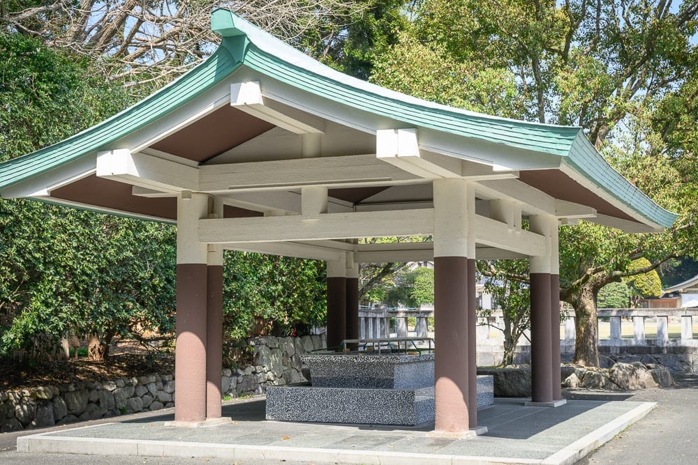 日本福岡-護國神社｜寺廟、涼亭、屋頂、石柱、水池、石牆、樹木，庭院戶外空間，綠瓦白牆亭子內部設有水槽，周圍環繞綠樹與石造