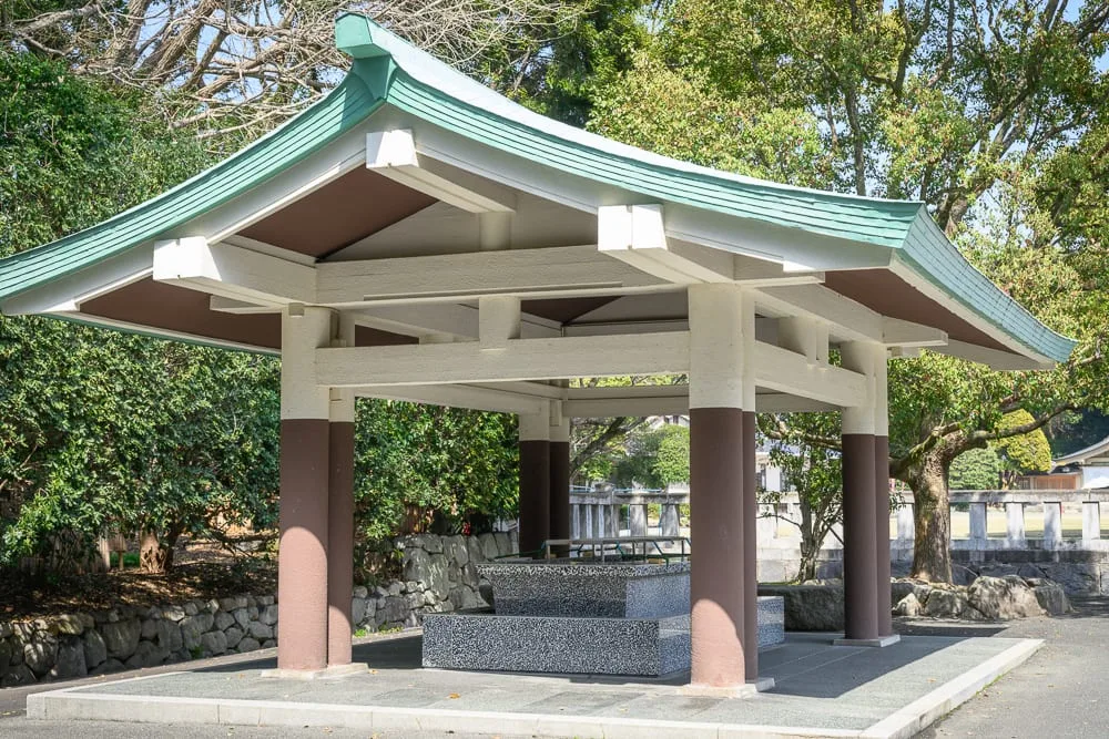 日本福岡縣護國神社完整攻略|御朱印收集與週末跳蚤市場 7 日本福岡-護國神社|寺廟、涼亭、屋頂、石柱、水池、石牆、樹木,庭院戶外空間,綠瓦白牆亭子內部設有水槽,周圍環繞綠樹與石造