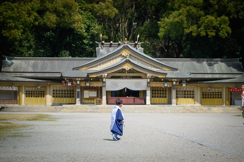 日本福岡-護國神社｜寺廟、孩童、廣場、樹木，身穿深藍和服的幼童站在灰色碎石廣場中央，背對鏡頭面向傳統日式神社正殿，後方為