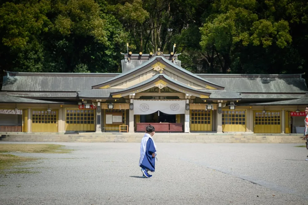 日本福岡縣護國神社完整攻略|御朱印收集與週末跳蚤市場 13 日本福岡-護國神社|寺廟、孩童、廣場、樹木,身穿深藍和服的幼童站在灰色碎石廣場中央,背對鏡頭面向傳統日式神社正殿,後方為