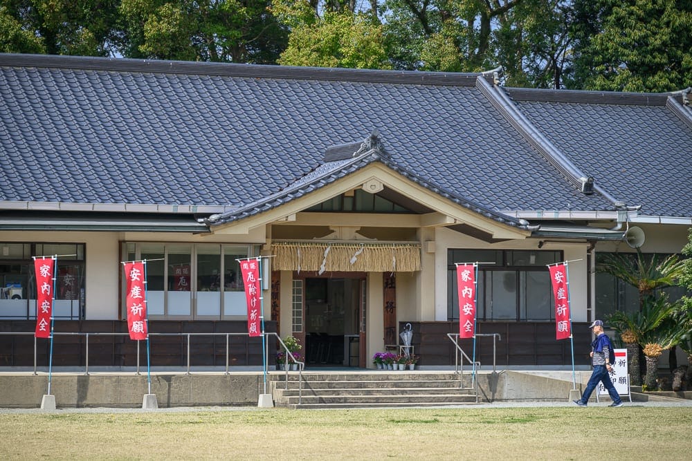 日本福岡-護國神社｜日式建築、瓦片屋頂、寺廟、旗幟、草地、行人，灰瓦黑梁的平房建築前方設有階梯入口，門口懸掛注連繩，數支