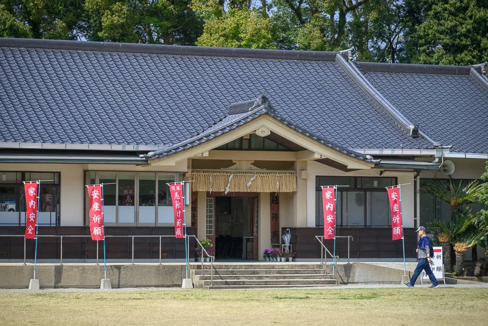 日本福岡縣護國神社完整攻略|御朱印收集與週末跳蚤市場 9 日本福岡-護國神社|日式建築、瓦片屋頂、寺廟、旗幟、草地、行人,灰瓦黑梁的平房建築前方設有階梯入口,門口懸掛注連繩,數支