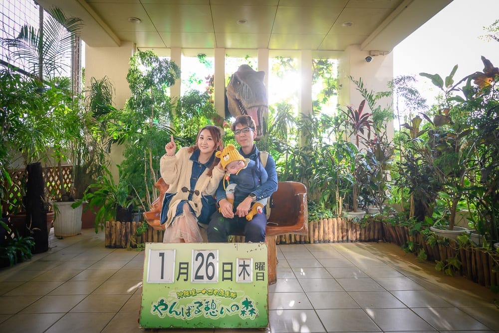 沖繩東濱恐龍公園完整攻略|親子免費景點玩法解析 7 日本沖繩恐龍公園|室內、木椅、人物、恐龍模型、盆栽植物,一家三口坐在鋪磁磚地板的長廊木椅上合影,前方立著標示「1月26