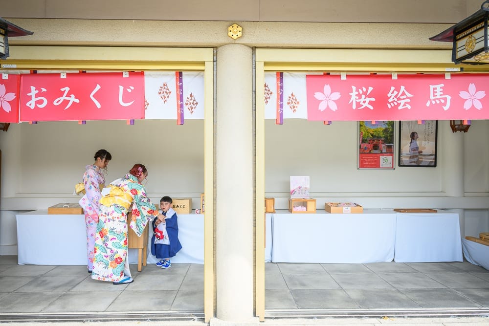 日本福岡-護國神社｜神社、御守販賣處、和服女子、兒童、橫幅布條、桌子、石地板，兩名穿著色彩鮮豔和服的女性與一名藍衣男童站
