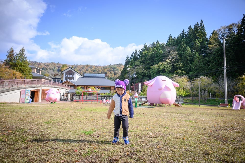日本福島-吉利蛋公園｜小孩，草地，粉紅吉祥物，橋，建物，樹林，藍天白雲。穿毛帽與外套的小孩站在草皮中央，後方散落粉紅胖丁造