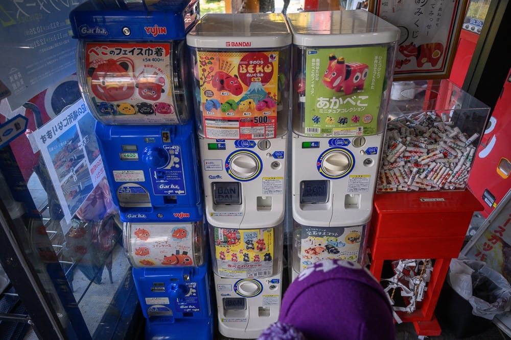 日本福島-吉利蛋公園｜室內、扭蛋機、紙條、收納箱，藍色與白色扭蛋機垂直堆疊並排，展示著紅牛玩偶圖樣廣告，右側紅色木架上放置