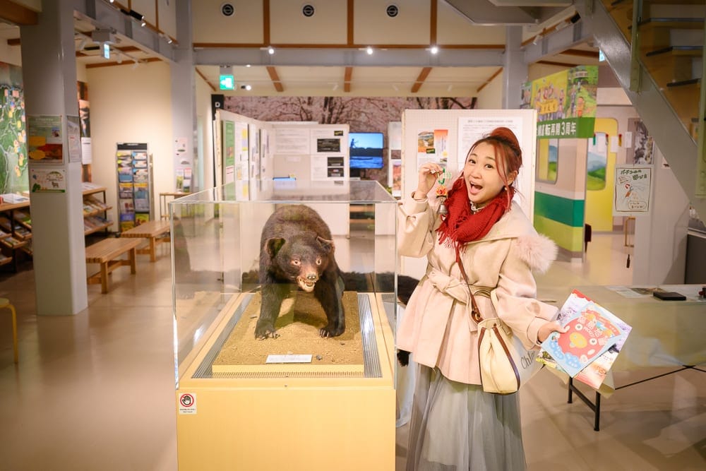 日本福島-吉利蛋公園｜室內、展覽館、熊標本、女子、展示櫃、宣傳冊、樓梯、立牌，明亮日光燈照明，女子身穿米色外套與紅圍巾，站