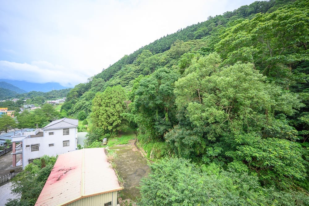 苗栗格拉斯行館｜山坡、綠樹、房屋、鐵皮屋頂、天空。山腳下坐落白色樓房與紅頂鐵皮屋，後方為一片綿延的茂密綠林，遠處可見