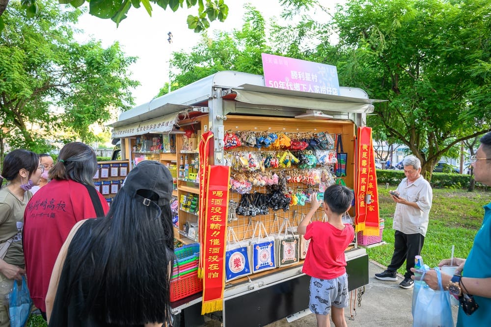 嘉義光織影舞｜戶外、小貨車、雜貨店、鑰匙圈、帆布袋、人群、綠樹。改裝小貨車兩側展示懷舊商品，吊掛鑰匙圈與帆布袋，多