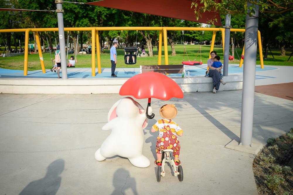 彰化田尾怡心園公園｜公園，黃色鞦韆架，白色兔子雕塑，小孩，腳踏車，遮陽傘，樹木，綠草地，大人，水泥地。