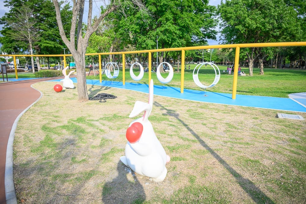 彰化田尾怡心園公園｜戶外公園，草地，白色兔子雕塑，紅色球體，黃色鐵架，白色圓形盪鞦韆，藍色跑道，樹木。
