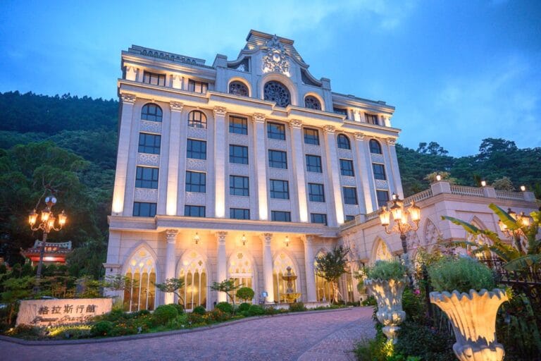 苗栗格拉斯行館 Grasse Grace Manor 住宿攻略|歐式城堡山林體驗完整指南 5 苗栗格拉斯行館|歐式城堡、階梯、路燈、植栽、山脈、黃色燈光。藍色暮色中,高聳的白色歐式建築外牆排列著方窗,前方有磚造
