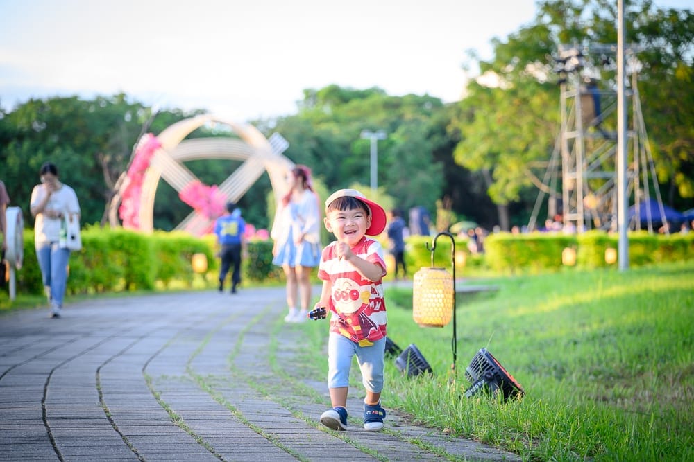 嘉義光織影舞｜草地、步道、小男孩、燈籠、樹木、路人。戴紅帽的小男孩在鋪磚步道上奔跑，旁邊草地上立著圓筒狀燈籠，背景