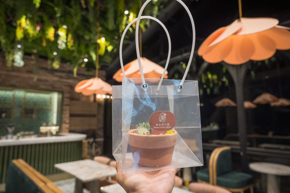 苗栗格拉斯行館｜室內，透明提袋，盆栽，花形吊燈，桌椅，吧台，綠植天花板。手持透明塑膠提袋裝著小型陶盆植物，背景為裝飾
