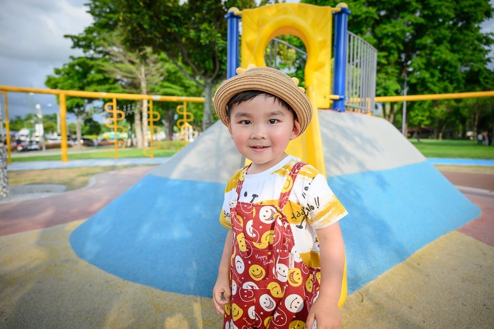 彰化田尾怡心園公園｜兒童，吊帶褲，草帽，溜滑梯，公園，樹木。戴著草帽的男孩站在戶外兒童遊樂場的彩色溜滑梯前方，身穿紅底笑
