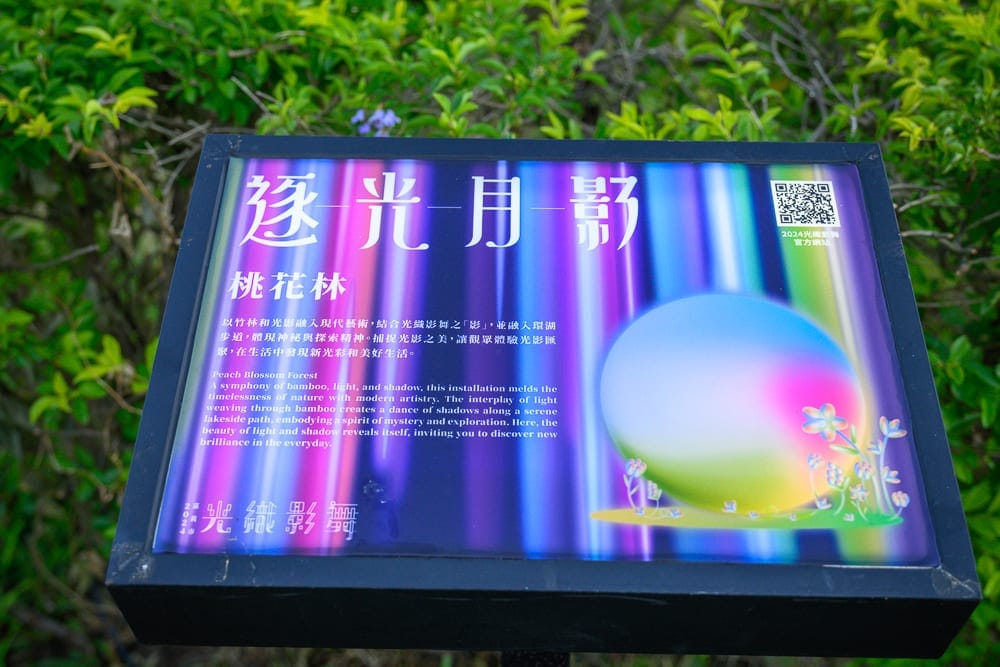嘉義光織影舞｜戶外綠叢中的金屬展示牌，牌面印著「逐光月影」、「桃花林」字樣與七彩光影圓球圖案，右上有二維碼。