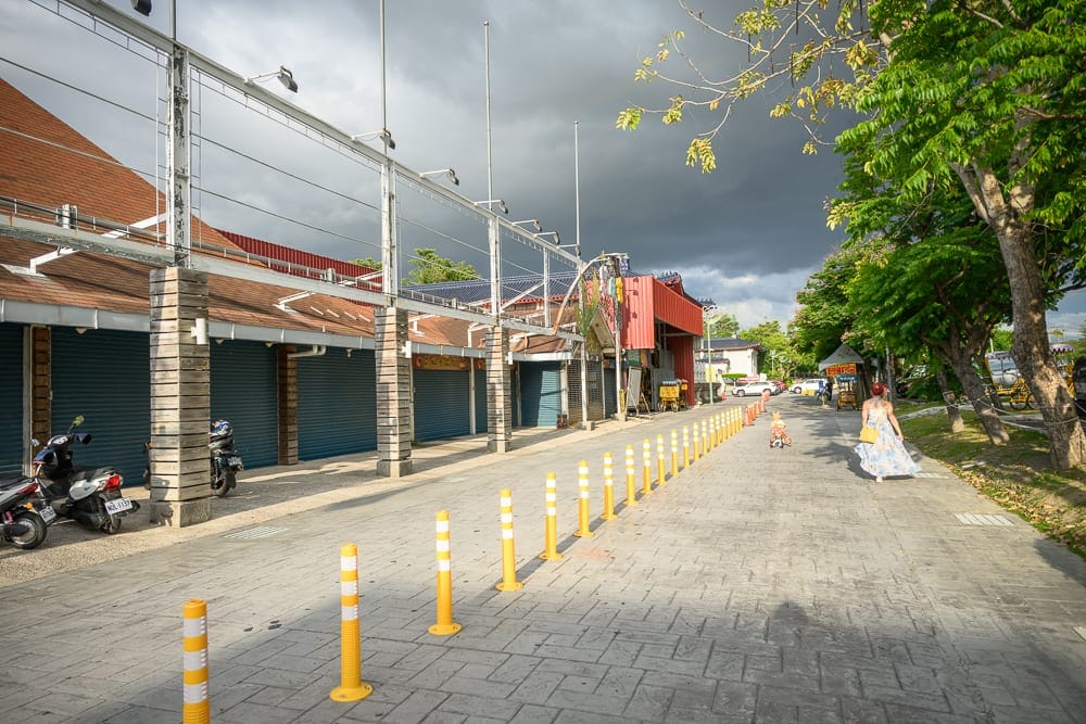 彰化田尾怡心園公園｜街道,店鋪,柱子,黃色塑膠柱,行人,機車,樹木。石板路旁為整排鐵捲門拉下的店鋪，前方立有黃色反光柱。
