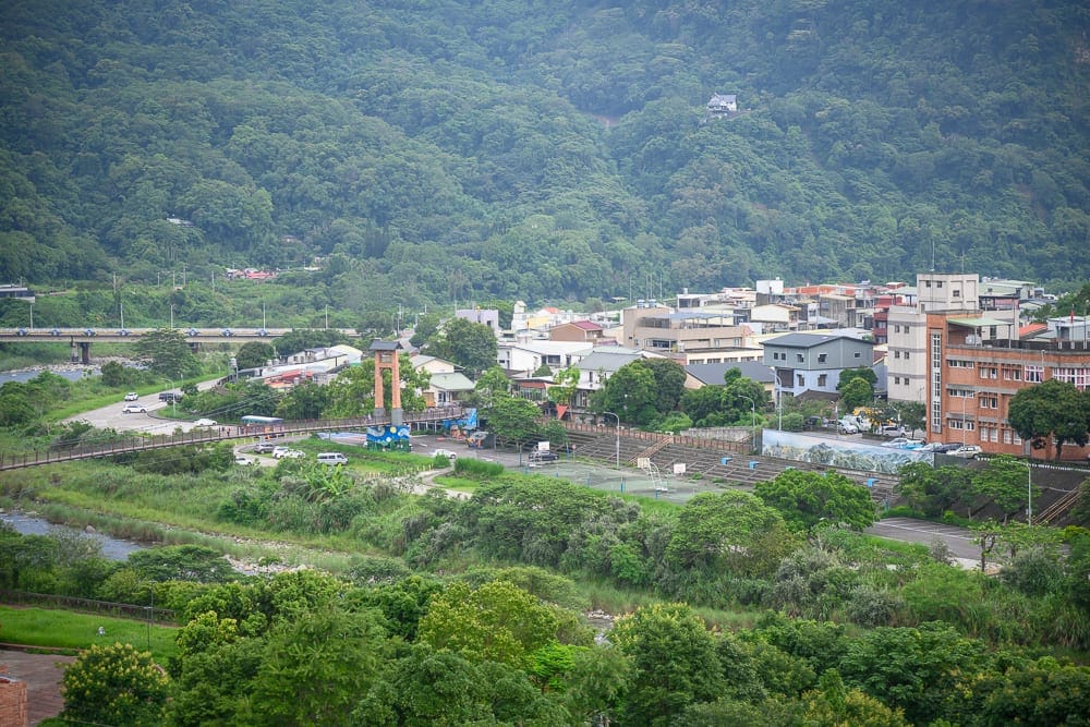 苗栗格拉斯行館｜山脈，河流，橋，建築，樹林，道路，車輛。層疊的綠意山頭下方有密集的低矮房舍，蜿蜒的河道伴隨帶狀植被，