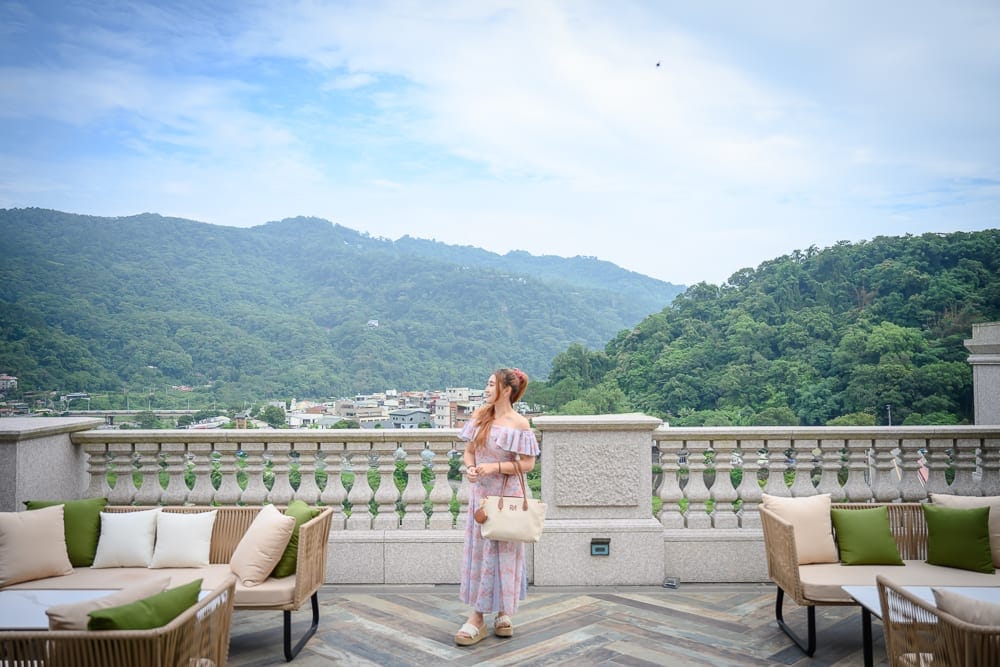 苗栗格拉斯行館｜女子、露台、沙發、山景、建築、天空。 露台上配置多張編織沙發與抱枕，一名女子手提包包站立於欄杆前。遠