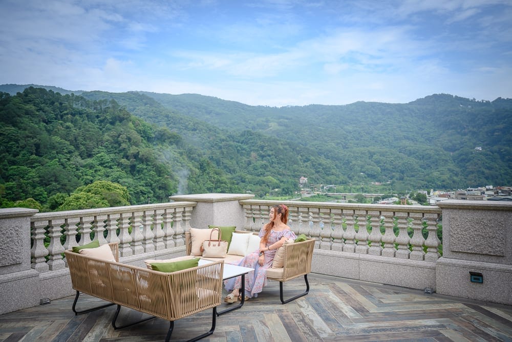 苗栗格拉斯行館｜露臺，沙發，抱枕，女性，欄杆，山景，天空。一名長髮女性穿著長裙坐在戶外露臺的編織沙發上，身旁擺放手提