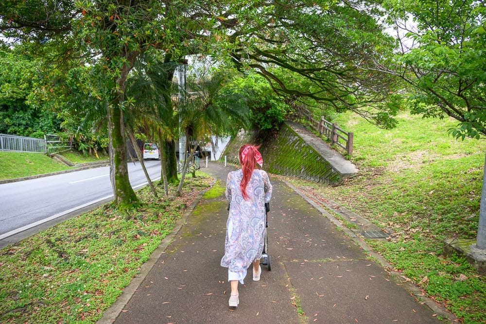 沖繩浦添大公園完整攻略|超長滾輪溜滑梯與親子遊樂設施 7 日本沖繩浦添大公園|女人、推車、人行道、馬路、大樹、護欄、草地,背對鏡頭的紅髮女子身穿花紋長衫,推著嬰兒車行走在綠樹成蔭