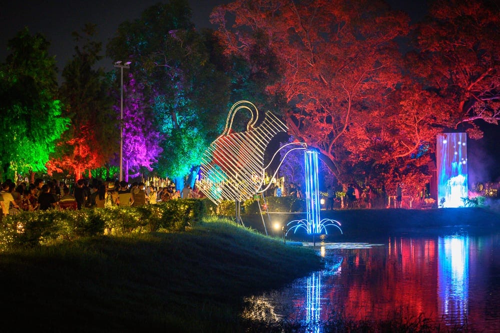 嘉義光織影舞｜夜晚的戶外公園水池畔，大型金屬水壺燈飾向池中傾倒出藍色線條的水流光影，後方樹林被紅、綠、紫燈光投射，