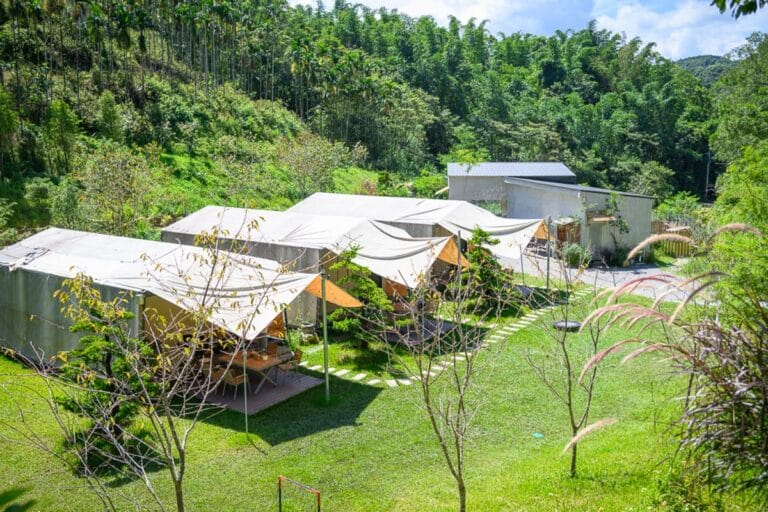 南投葉子秘境｜草地，露營帳篷，遮陽帆布，樹木，建築，山林，戶外。幾座連棟白色帳篷座落於綠色草地上，邊緣擺放桌椅，周