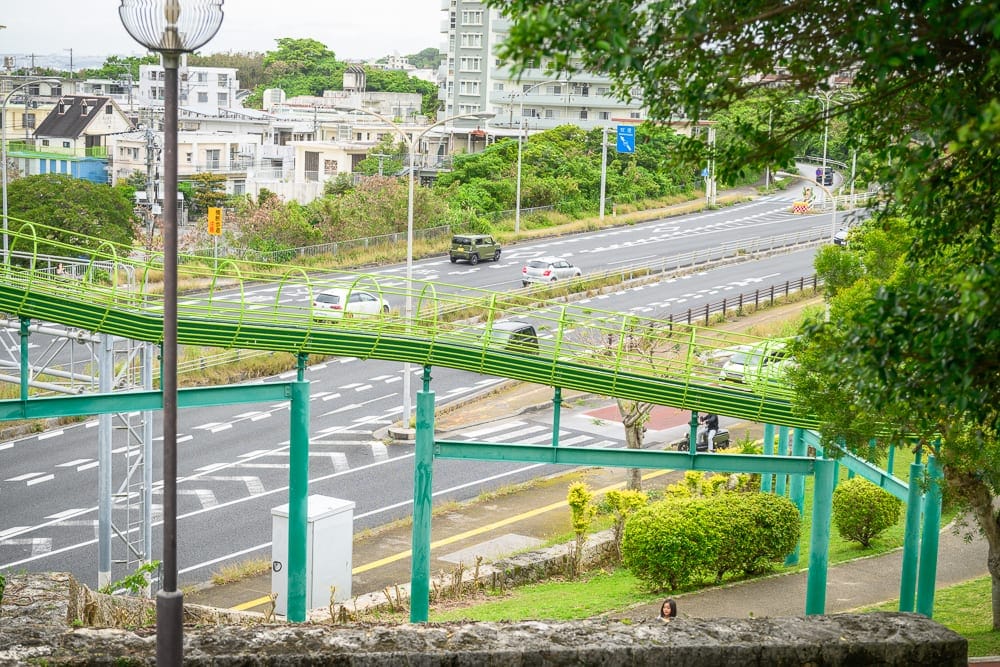 沖繩浦添大公園完整攻略|超長滾輪溜滑梯與親子遊樂設施 8 日本沖繩浦添大公園|綠色管狀天橋、多車道馬路、遠處住宅區、路燈桿、綠樹、灰色石牆,天橋橫跨車流量平穩的寬敞柏油路面,周邊