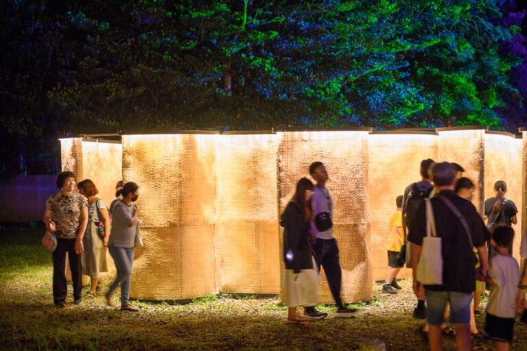 嘉義光織影舞完整攻略|北香湖公園夜間裝置藝術與市集玩法解析 10 嘉義光織影舞|夜間戶外草地,行人,發光竹編圓柱裝置藝術,綠樹,燈光投射。人們穿梭在環形發光柱體周圍,背景為被藍綠色