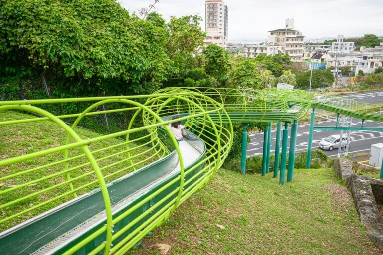 沖繩浦添大公園完整攻略|超長滾輪溜滑梯與親子遊樂設施 1 日本沖繩浦添大公園|綠色鋼管溜滑梯、草坡、孩童、高架道路、遠方房舍。孩童正坐在長型溜滑梯中往下滑行,滑梯外側包覆著圓形的