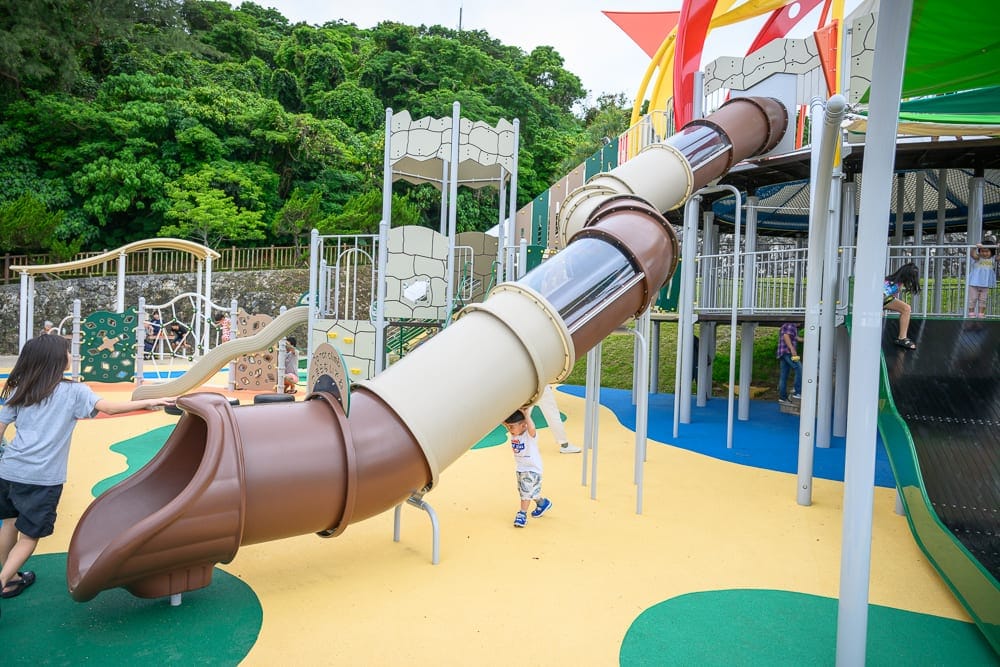 沖繩浦添大公園完整攻略|超長滾輪溜滑梯與親子遊樂設施 13 日本沖繩浦添大公園|戶外遊樂場,透明管狀滑梯,兒童,彩色地面,攀爬架,綠色樹林。